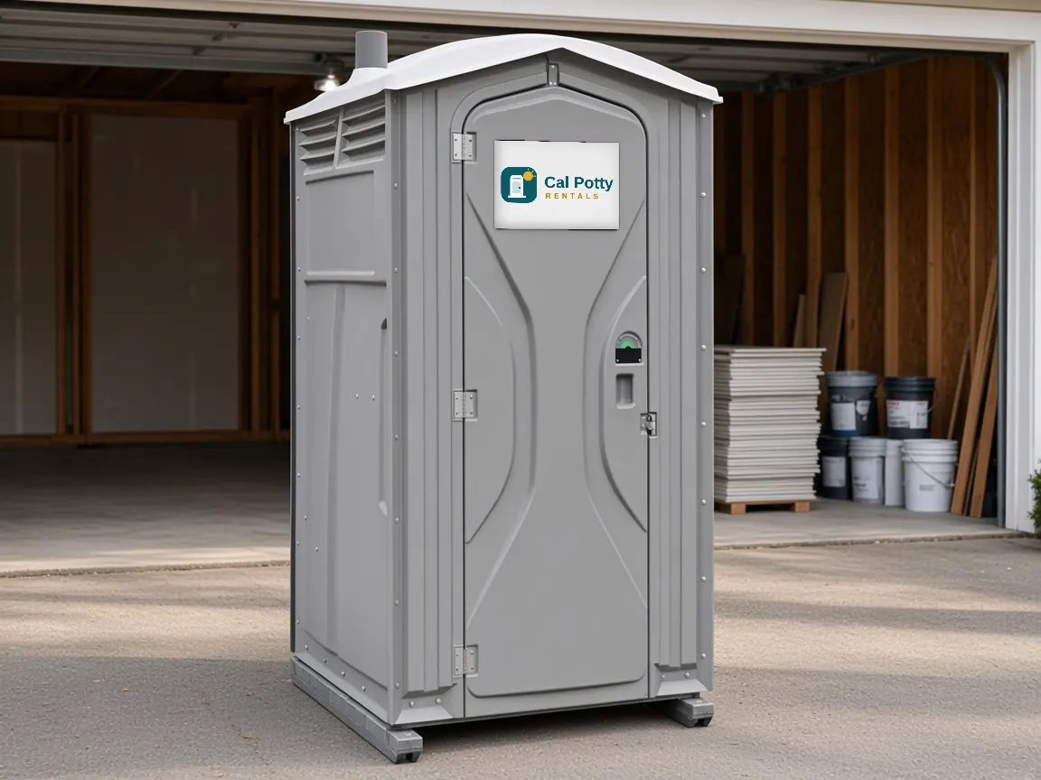 A grey porta potty at the open garage of a home with paint buckets and lumber